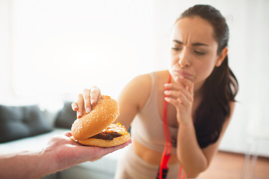 Young woman doing yoga workout in room during quarantine. Girl with red measure tape across shoulder hold upper part of burger and think. Want to eat or not.
