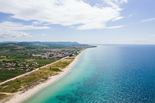 Aerial view of the long sandy beach and turquoise Aegean Sea waters bordering the coastal village and green fields in Paralia Ofriniou, Greece.