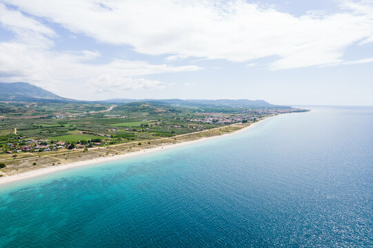 Aerial view of the sandy coastline and turquoise waters with green fields and distant mountains in Paralia Ofriniou, Greece.