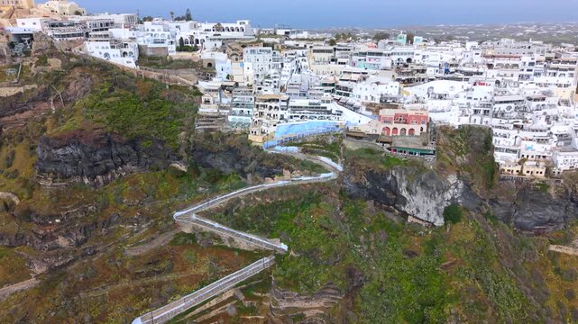 A dramatic high-angle aerial video of the famous zigzagging stone staircase in Fira, Santorini, descending the steep volcanic cliffs from the white village down toward the Aegean Sea.