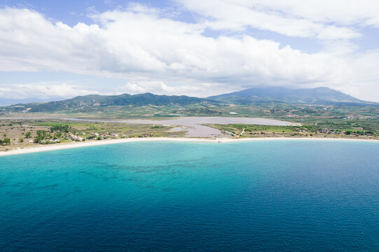 Aerial view of the turquoise coastline and sandy beach with a coastal lagoon and green hills under a cloudy sky in Paralia Ofriniou, Greece.