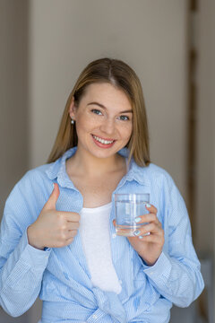 A cheerful woman in a casual outfit holds a glass of water and gestures a thumb up