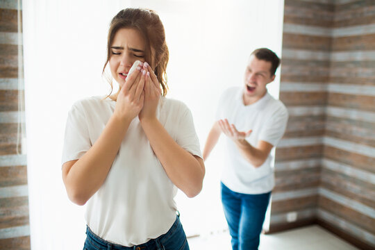 Young woman and man in room during quarantine. Guy scream at his wife. She crying and hold white tissue on cheek. Abusing and home agresiveness.