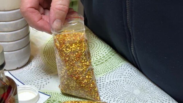 Close-up seller holding transparent pollen bag hand tamping golden granules into sealed gram pouch on patterned tablecloth indoor