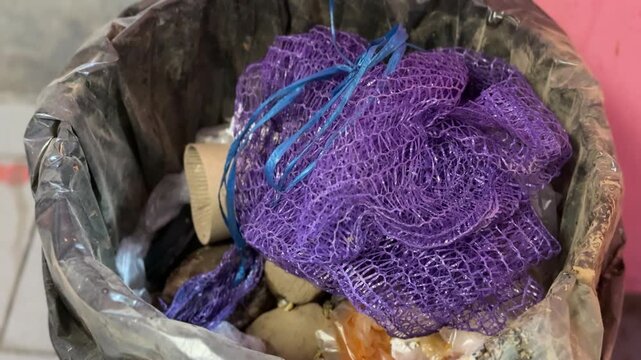 Close-up green trash can overflowing with purple cloth and scrubber, plastic liner packed with food scraps and crumpled packaging