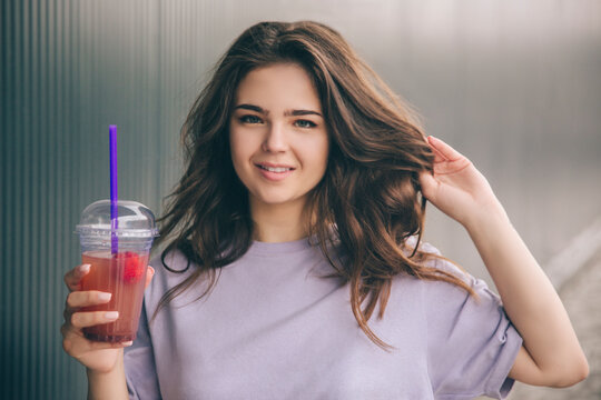 Young stylish trendy woman isolated over grey blue background. Beautiful attractive girl posing on camera. Holding pne hand in hair and cup of lemonade with another one.