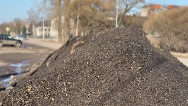 Dirty snow pile along highway under blue sky, mud and gravel mixed into melting slush, parked cars and bare trees in background