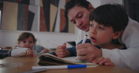 Naklejka premium Mother helping son concentrate on drawing in sketchbook while baby sibling reaches curiously from high chair, portraying everyday family life and bonding at home