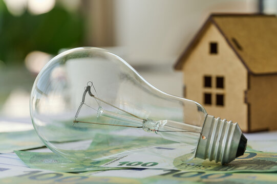 A light bulb rests on banknotes next to a model house. The scene shows concepts of money management and housing.