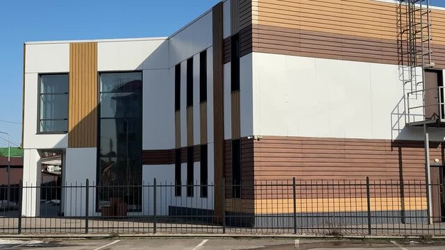 Stylish modern house with siding under blue sky, geometric facade of glass and wood panels, parking lot and metal fence