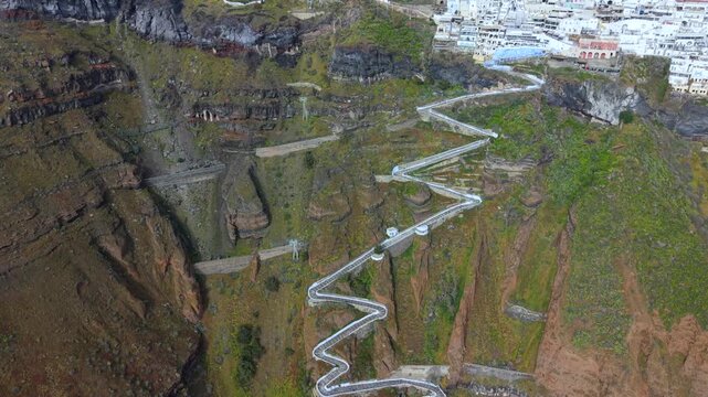 A dramatic high-angle aerial video of the famous zigzagging stone staircase in Fira, Santorini, descending the steep volcanic cliffs from the white village down toward the Aegean Sea.