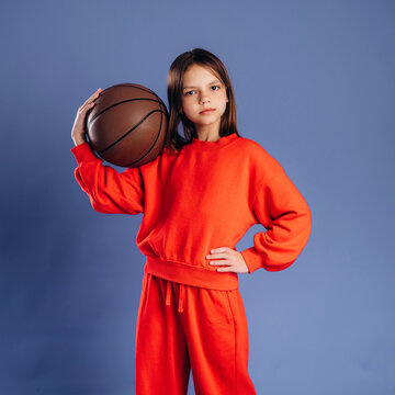 Young girl wearing bright red sportswear holds a basketball in one hand while standing confidently against a blue background