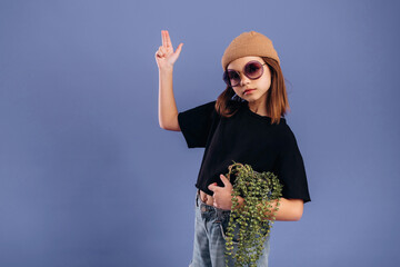 Fototapeta premium Young girl wearing sunglasses and a beanie poses confidently with a plant in her hand against a solid purple background, showcasing a trendy urban style