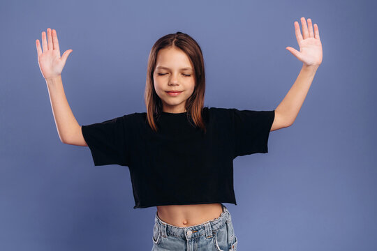 Young girl with brown hair wearing a black crop top and denim shorts raises her arms in a relaxed pose against a solid blue background
