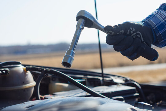 Gloved hand holding a socket wrench with extension over an open car engine bay. Concept vehicle diagnostics, roadside assistance, DIY auto repair and professional mechanic services.