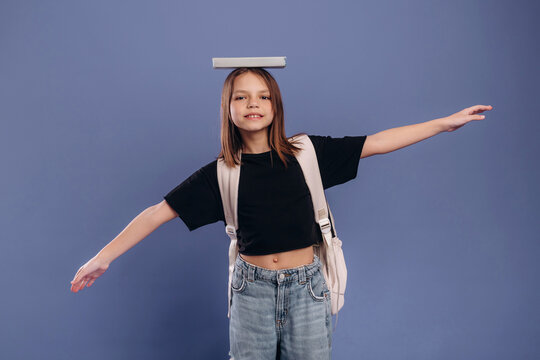 Young girl balancing a book on her head while standing with arms outstretched, wearing a black t-shirt and denim jeans against a solid blue background