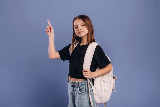 Young girl with long brown hair wearing a black shirt and blue jeans holds a white backpack and points upwards against a solid blue background