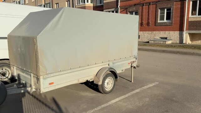 Covered cargo trailer parked lot spring, pale tarp stretched over metal frame, single axle wheel visible, sunlit asphalt surface