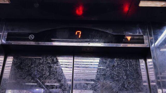 a metal elevator display showing number seven and a red downward arrow. The lift is moving from the seventh floor to ground level inside a dimly lit building interior.