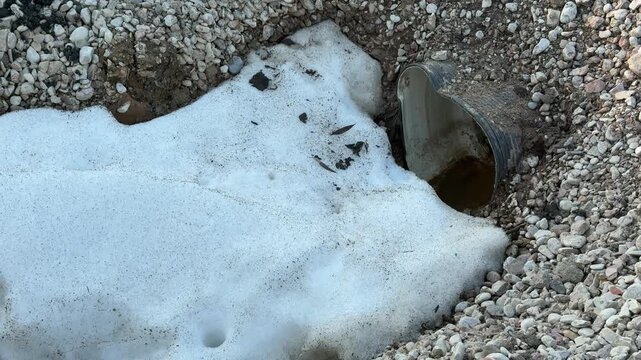 Snow on crushed stone around drain, concrete rim, asphalt edge, small sewer pipe opening with melting runoff and silt, pale winter