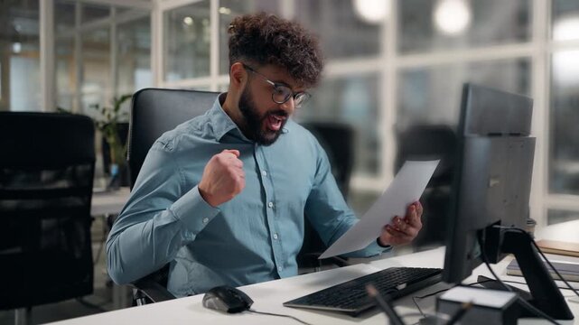 Ethnic Arabian business man working computer win holding papers report happy rejoice good result financial success smiling businessman looking at desktop hold document work winning male guy at office