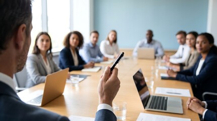 Fototapeta premium Business leader standing at head of conference room table holding pen while gesturing during team meeting with colleagues working on laptops in modern bright corporate boardroom setting