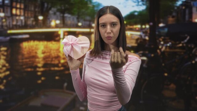 Young woman holding heart shaped gift and beckoning with open hand on street bridge at night; romantic invitation.