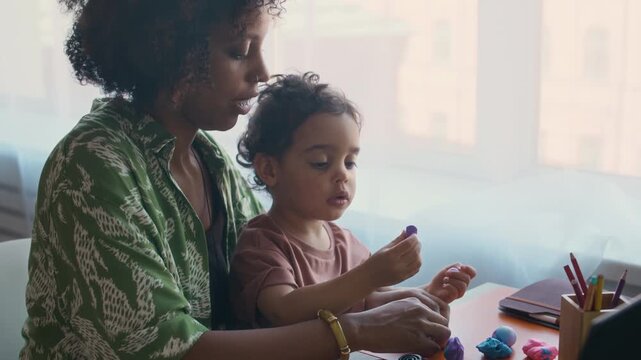 Zoom in handheld shot of young kid sitting on his mothers lap while modeling with clay together at home table