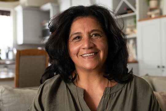 Mature woman sitting on sofa at home, wearing olive-green blouse with thin gold necklaces, cabinets