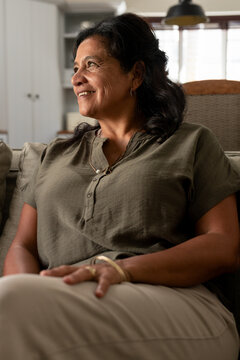 African American woman sitting on sofa in living room, wearing olive top, necklace rings lamp