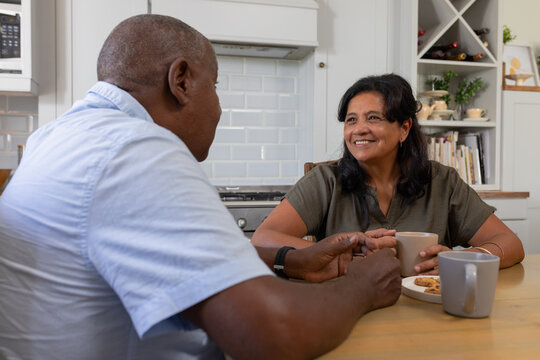 Mature African American couple holding mugs, sitting at wood table in kitchen with plate of cookies