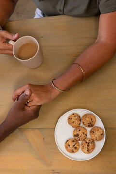 Senior couple sitting at home holding hands sharing coffee from mug with cookie plate and ring