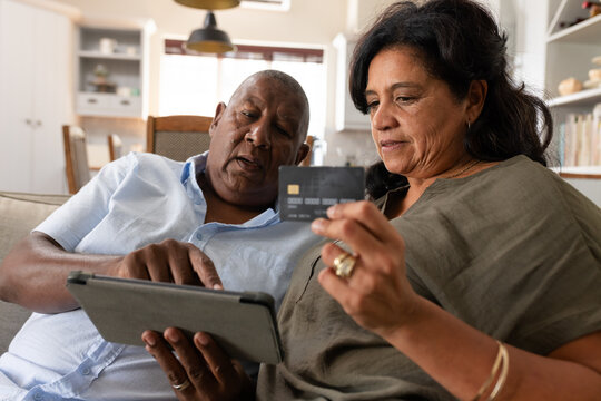 Senior couple sitting on sofa in living room, man using tablet while woman holding credit card