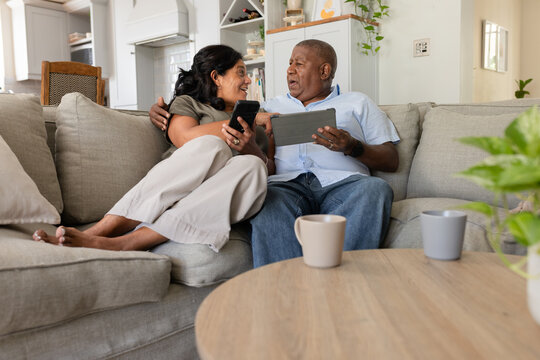 Senior couple sitting closely on light-gray sectional sofa at home holding smartphone and tablet