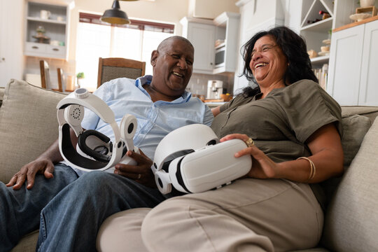 African American couple laughing, sitting on light sofa in open kitchen, holding white VR headsets