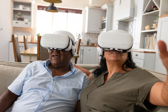 African American senior couple wearing white VR headsets sitting on sofa at home reaching outward