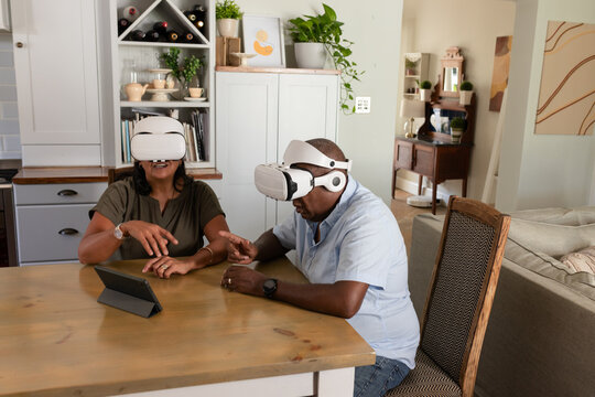 African American couple wearing white VR headsets and interacting with tablet at home dining table