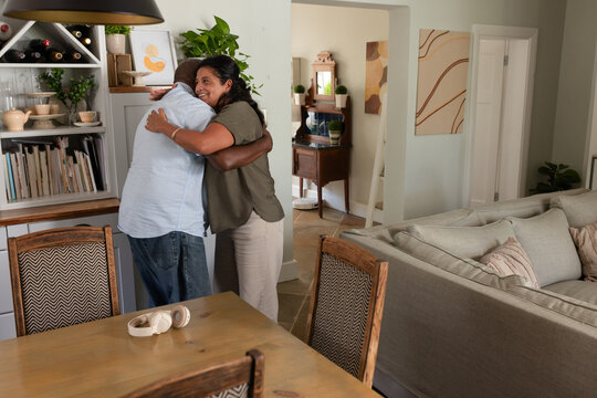 African American couple hugging, smiling by shelving in home dining area with headphones on table