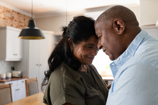 Senior couple pressing foreheads and smiling in kitchen at wooden dining table under pendant light