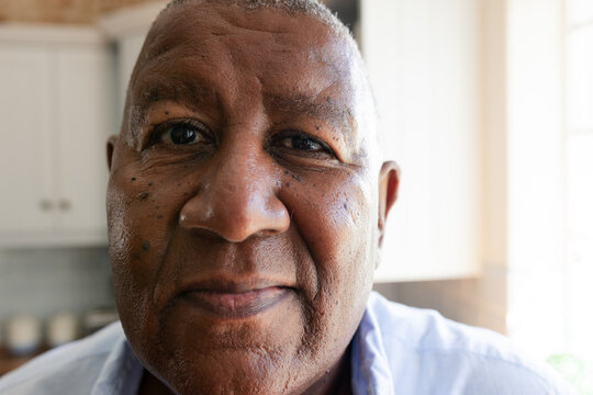 Senior African American man wearing light button-up shirt, gazing into camera by window in kitchen