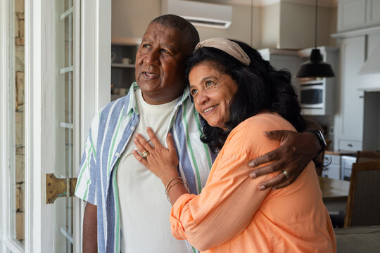 African American senior couple standing embracing in kitchen by glass door, showing rings and watch