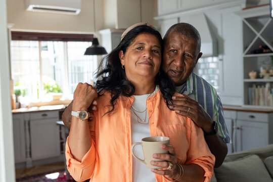 African American couple standing in kitchen, woman in coral shirt holding beige mug, man leaning