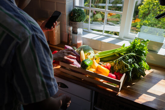 Mature man holding smartphone and reaching for vegetables in wooden crate on sunlit counter