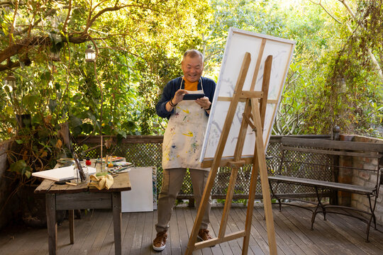 Senior Asian man standing at wooden easel holding tablet, wearing paint apron on garden terrace