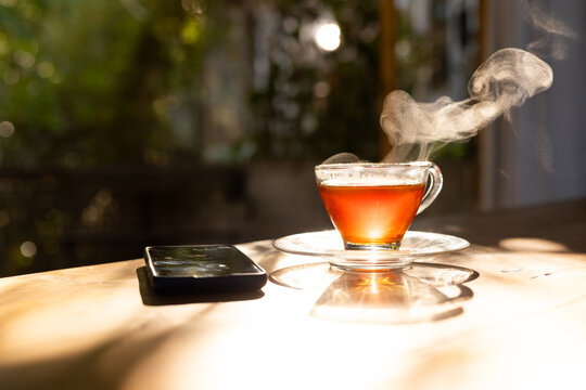 Clear glass teacup holding steaming amber tea on sunlit table phone face-up left blurred foliage