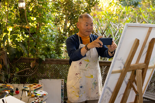 Senior Asian man painting on wooden deck with easel, wearing painted apron, checking smartphone