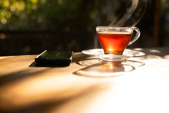Clear glass teacup is steaming on wooden patio table while black smartphone is resting, copy space