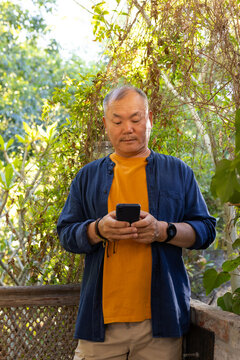 Senior Asian man wearing orange tee and blue shirt standing on porch checking smartphone by trellis