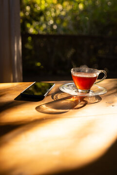 Glass teacup is steaming on wooden table while smartphone is reflecting green foliage outside