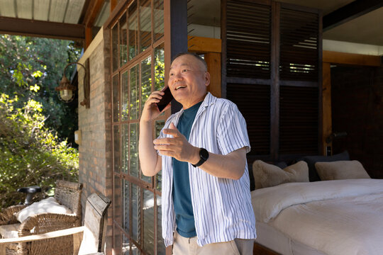 Senior Asian man standing in bedroom doorway to patio, smiling, speaking on phone, holding glass